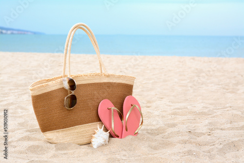 Stylish straw bag with sunglasses, flip flops and shell on sand near sea, space for text. Beach accessories
