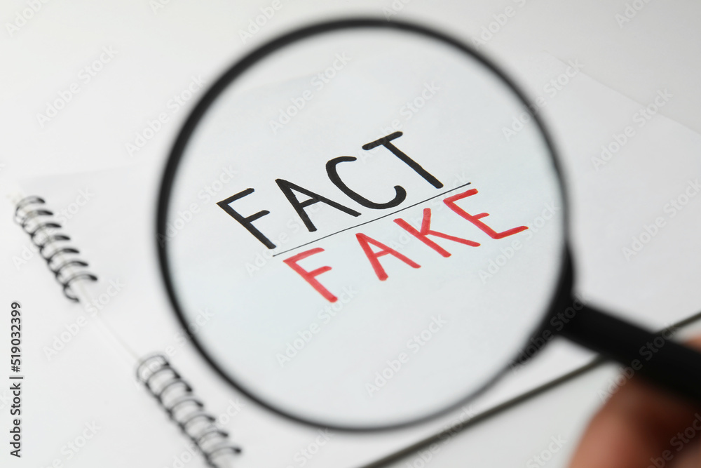 Woman holding magnifying glass over notebook with words Fact and Fake on white background, closeup Woman holding magnifying glass over notebook with words Fact and Fake on white background, closeup