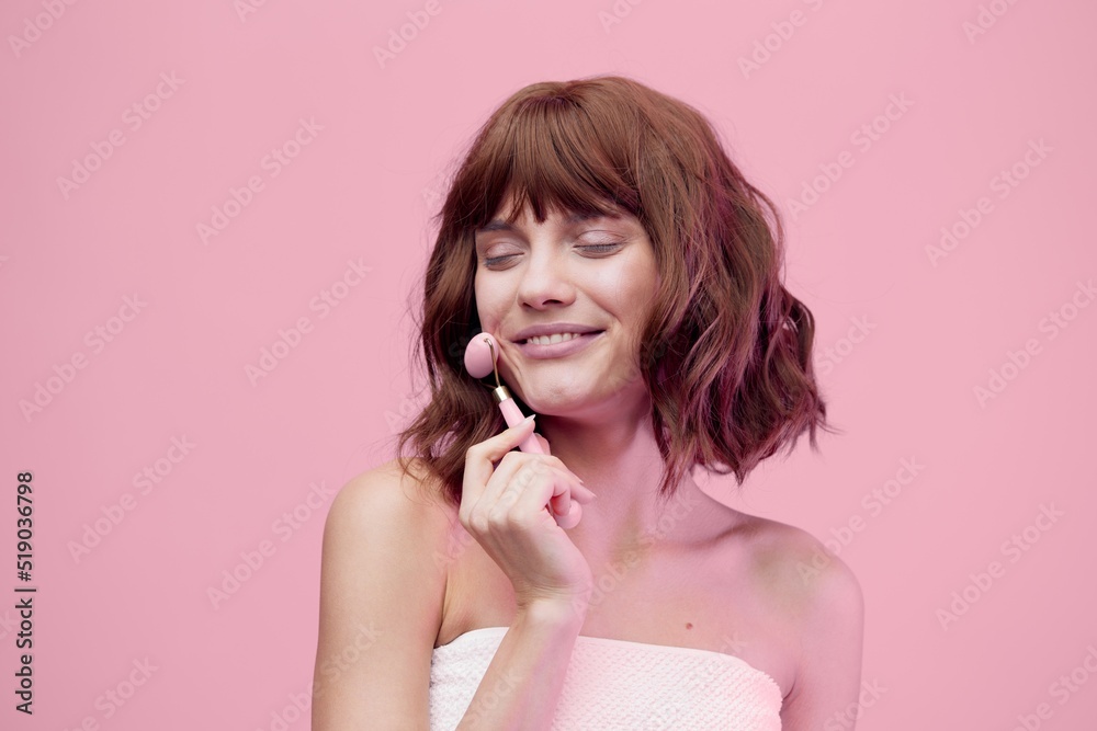 happy woman standing on a pink background with pink lighting from the side, with beautiful, wave-styled hair, closing her eyes with pleasure while doing a facial massage with a pink roller happy woman standing on a pink background with pink lighting from the side, with beautiful, wave-styled hair, closing her eyes with pleasure while doing a facial massage with a pink roller