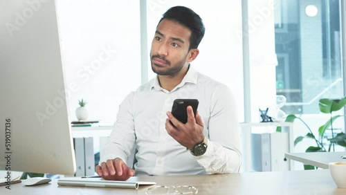 Young business man reading a message on his phone and working on his computer in the office. Handsome corporate professional sending an email and updating the project deadline after receiving a text