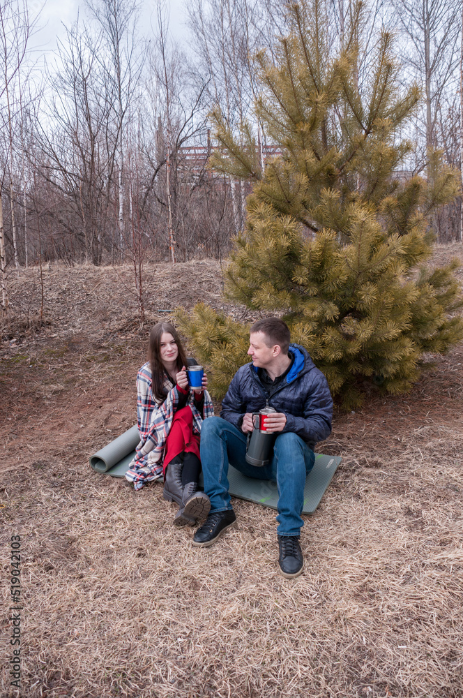 A couple in a spring park: a man and a girl sit near a fir tree and drink hot tea from a thermos A couple in a spring park: a man and a girl sit near a fir tree and drink hot tea from a thermos