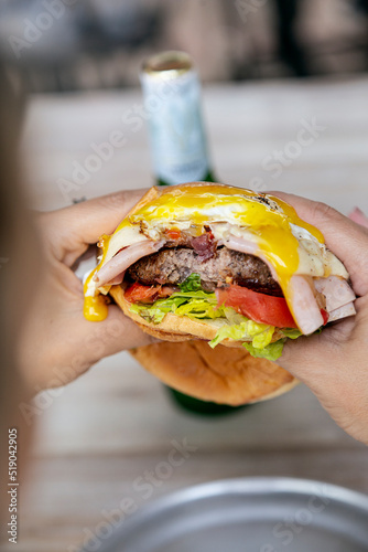 Cheeseburger with ham, fried egg, lettuce, tomato and onion, bitten by a person holding it in his hands.