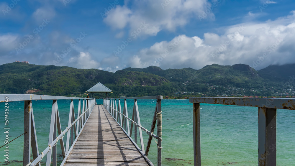 A wooden pedestrian walkway with metal railings runs over the turquoise ocean. There is a canopy in front. Green mountains of the island against a blue sky with clouds. Seychelles