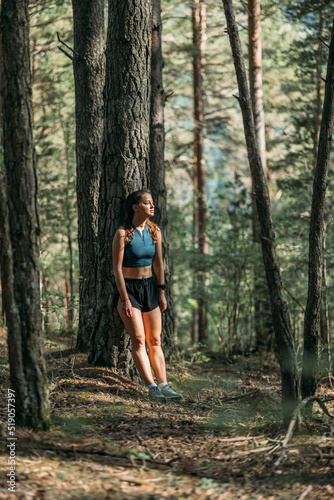 Tired young woman in sportswear leaned on a tree and resting in the forest after working out. Trail running and active life concept.