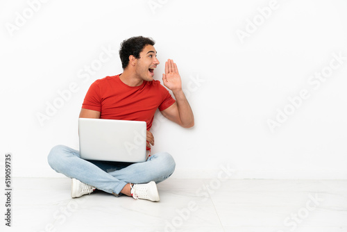 Caucasian handsome man with a laptop sitting on the floor shouting with mouth wide open to the lateral