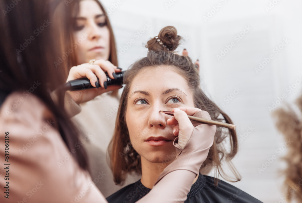 Fototapeta premium Professional make-up and hair style artists doing make-up and hair curls to a beautiful caucasian woman in a beauty salon
