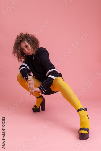 Stylish, bright, emotional cool style young woman dressed in yellow tights, sweater. Fashion Girl with curly hair and colored make-up posing on a pink background