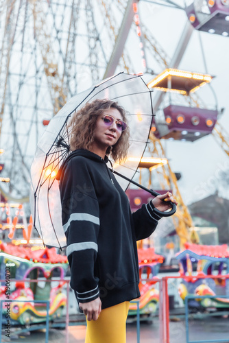Portrait of a young stylish woman with curly hair and colored make-up posing with transparent umbrella in amusement park. Street fashion