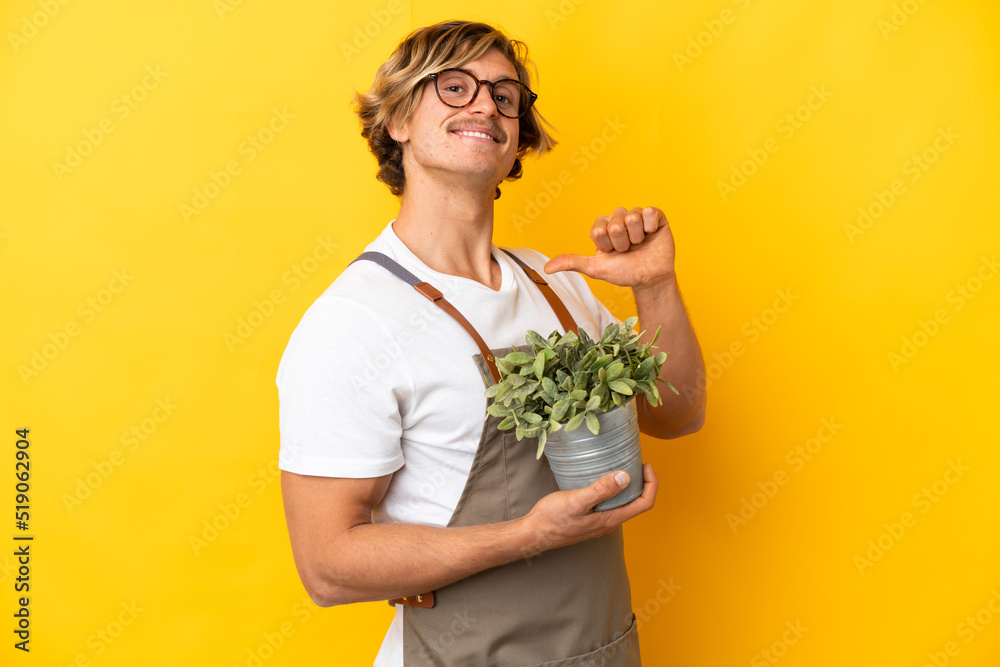 Gardener blonde man holding a plant isolated on yellow background proud and self-satisfied