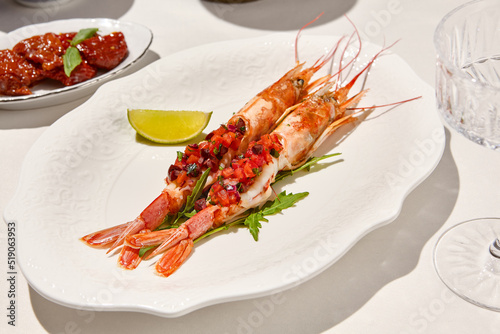 Seafood appetizer - baked langoustines with tomato tartar on white table. Grilled shrimp in italian restaurant menu. Langoustines on white background with shadows. Shrimp appetizer. Baked prawn.