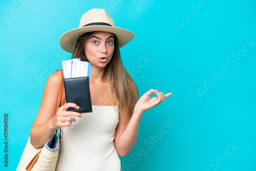 Young woman in swimsuit holding passport isolated on blue background surprised and pointing side