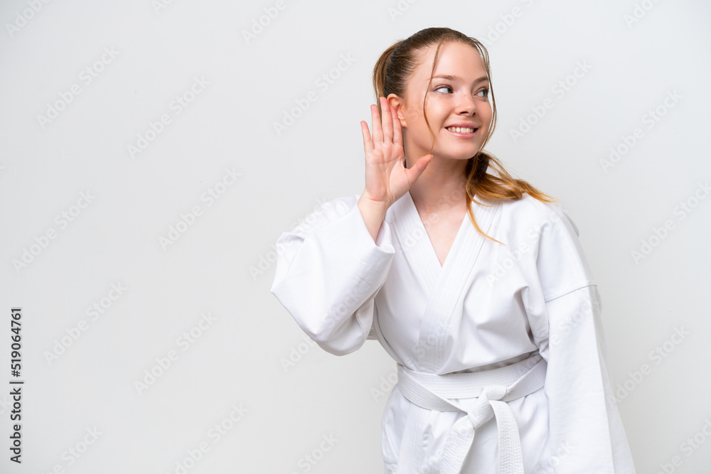 Young caucasian girl doing karate isolated on white background listening to something by putting hand on the ear Young caucasian girl doing karate isolated on white background listening to something by putting hand on the ear