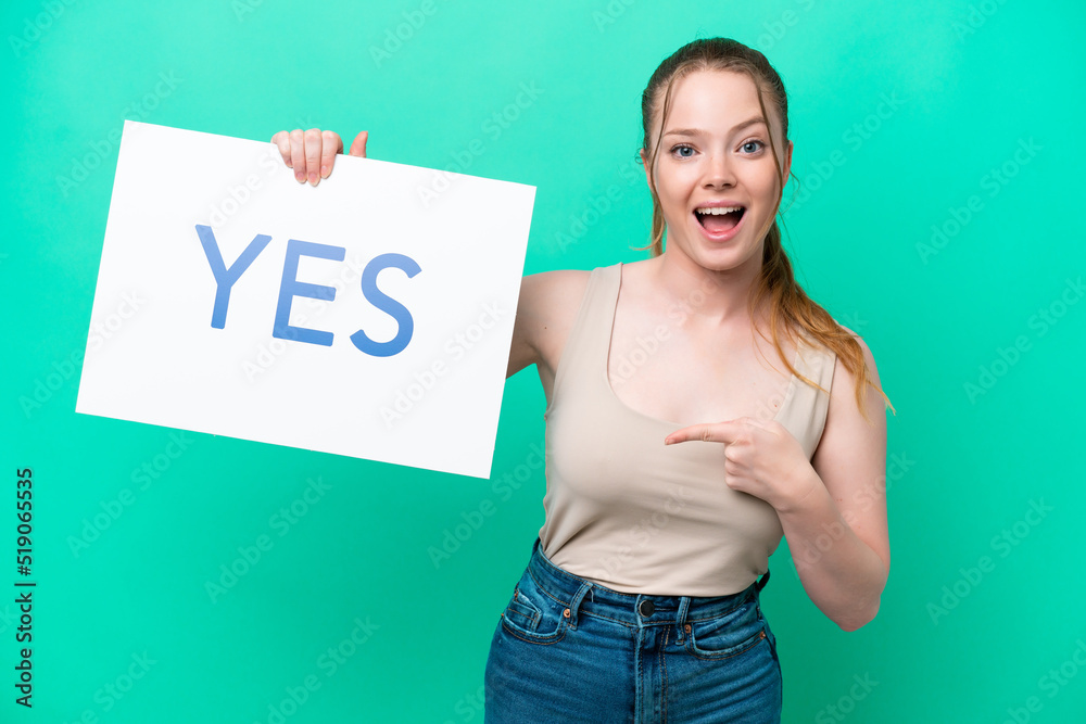 Young caucasian woman isolated on green background holding a placard with text YES and pointing it Young caucasian woman isolated on green background holding a placard with text YES and pointing it