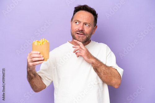 Middle age caucasian man holding fried chips isolated on purple bakcground looking up while smiling © luismolinero