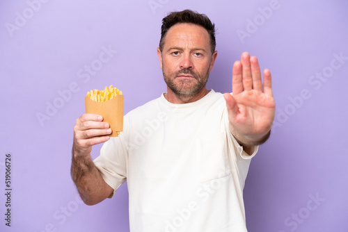 Middle age caucasian man holding fried chips isolated on purple bakcground making stop gesture © luismolinero
