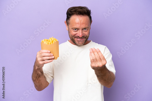 Middle age caucasian man holding fried chips isolated on purple bakcground inviting to come with hand. Happy that you came © luismolinero