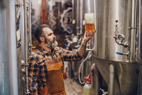 Bearded brewery master holding glass of beer and evaluating its visual characteristics. Small family business, production of craft beer. photo