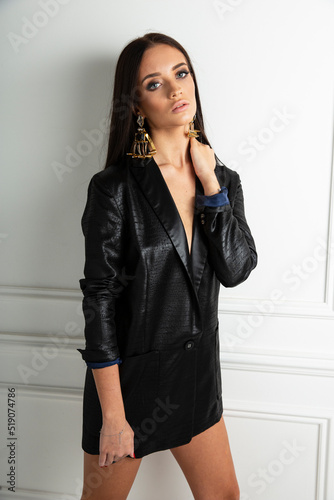 A brunette young woman with makeup dressed in leather black blazer and masive earrings, posing near white wall. photo