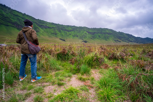 Photo of backside of asian man standing in the middle of meadow in mount bromo on a cloudy morning