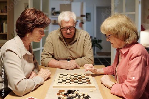Serious aged woman thinking of where to move checker while sitting by table with chessboard among her friends during leisure game