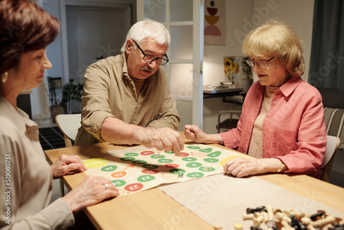 Senior man moving chip throughout paper board with green and red circles while playing leisure game with two mature women