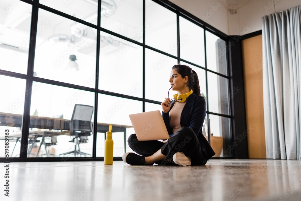 Thoughtful businesswoman sitting on ground with laptop and water bottle in office Thoughtful businesswoman sitting on ground with laptop and water bottle in office