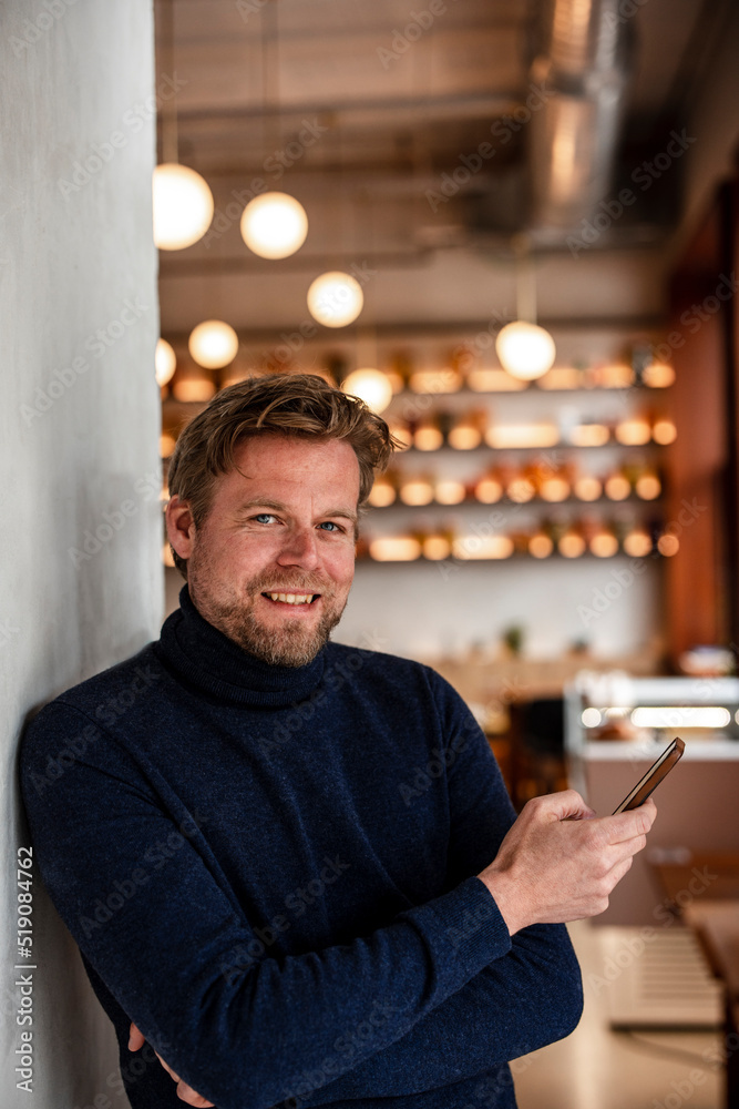 Smiling businessman with mobile phone leaning on wall in cafe Smiling businessman with mobile phone leaning on wall in cafe