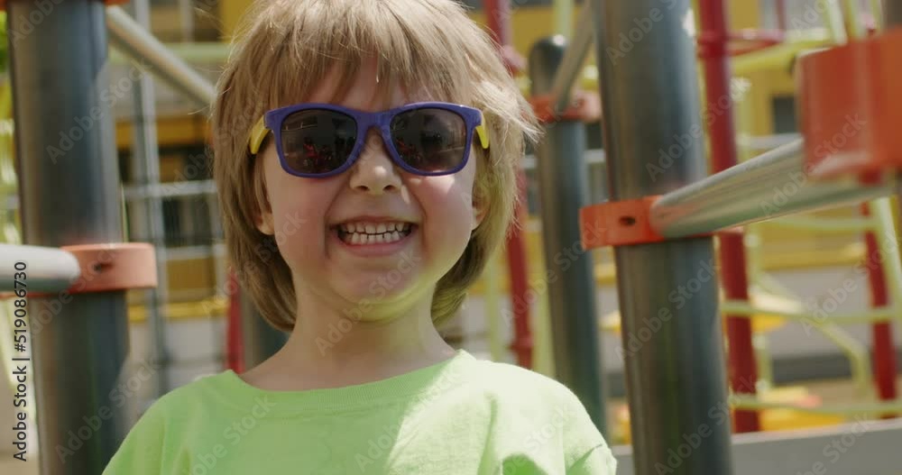 Blond boy in sunglasses and green T-shirt on children playground joyfully laughs at camera. Little kid has fun on playground on summer day closeup Blond boy in sunglasses and green T-shirt on children playground joyfully laughs at camera. Little kid has fun on playground on summer day closeup