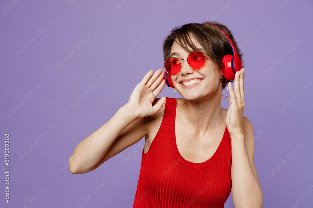 Young happy dreamful smiling fun woman 20s she wear red tank shirt eyeglasses headphones listen to music dance look aside isolated on plain purple backround studio portrait People lifestyle concept. Young happy dreamful smiling fun woman 20s she wear red tank shirt eyeglasses headphones listen to music dance look aside isolated on plain purple backround studio portrait People lifestyle concept.