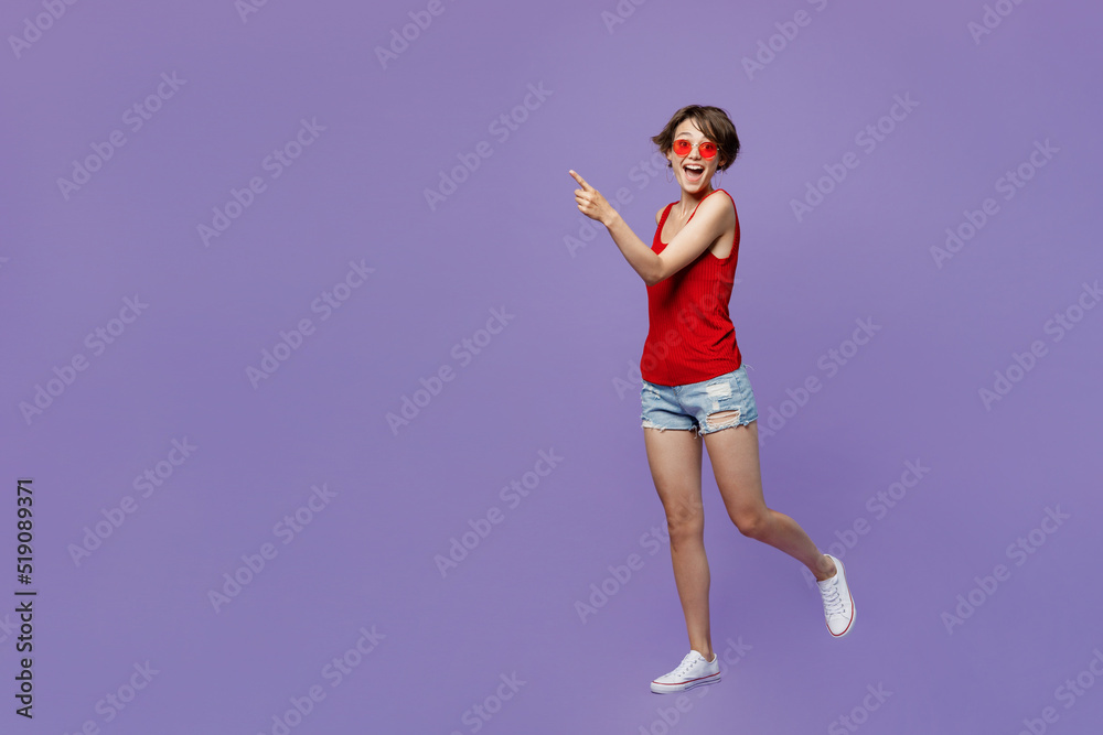 Full body young smiling happy fun woman 20s she wear red tank shirt eyeglasses walking goign strolling point index finger aside on workspace area isolated on plain purple backround studio portrait Full body young smiling happy fun woman 20s she wear red tank shirt eyeglasses walking goign strolling point index finger aside on workspace area isolated on plain purple backround studio portrait