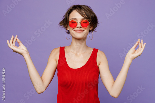 Young spiritual woman 20s she wear red tank shirt eyeglasses hold spreading hands in yoga om aum gesture relax meditate try to calm down isolated on plain purple backround. People lifestyle concept.