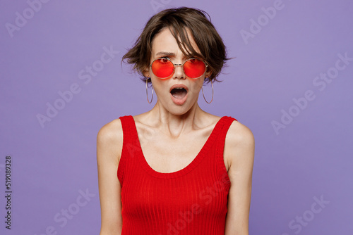 Young shocked angry annoyed mad sad furious woman 20s she wearing red tank shirt eyeglasses look camera with open mouth isolated on plain purple backround studio portrait. People lifestyle concept