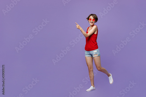 Full body young smiling happy fun woman 20s she wear red tank shirt eyeglasses walking goign strolling point index finger aside on workspace area isolated on plain purple backround studio portrait © ViDi Studio