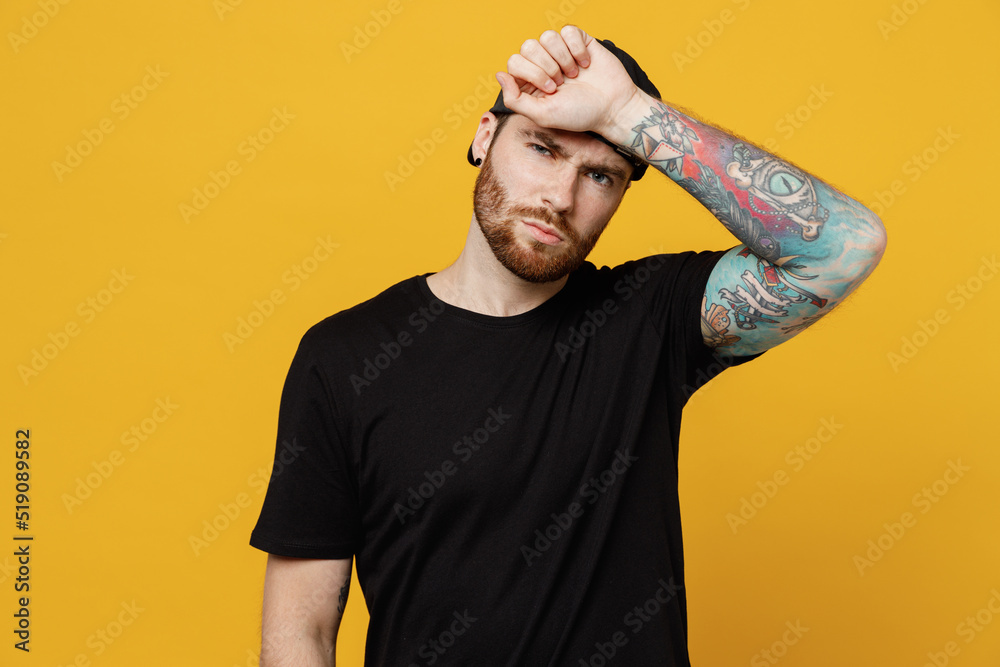 Young sick ill bearded tattooed man 20s he wears casual black t-shirt cap put hand on forehead suffer from headache solated on plain yellow wall background studio portrait. People lifestyle concept. Young sick ill bearded tattooed man 20s he wears casual black t-shirt cap put hand on forehead suffer from headache solated on plain yellow wall background studio portrait. People lifestyle concept.