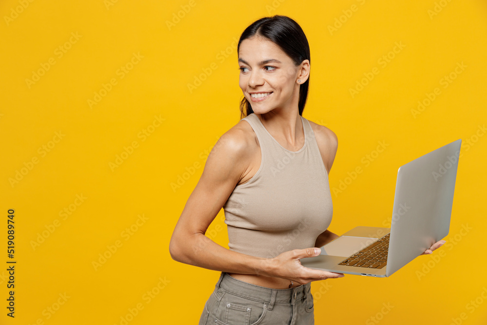 Naklejka premium Side view young smiling cheerful fun cool happy latin woman 30s she wearing basic beige tank shirt hold use work on laptop pc computer look aside on workspace area isolated on plain yellow backround
