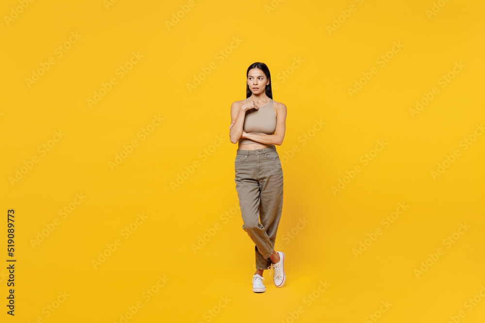Full body serious calm beautiful young latin woman 30s she wearing basic beige tank shirt look camera look aside on worksapce area mock up isolated on plain yellow backround People lifestyle concept. Full body serious calm beautiful young latin woman 30s she wearing basic beige tank shirt look camera look aside on worksapce area mock up isolated on plain yellow backround People lifestyle concept.