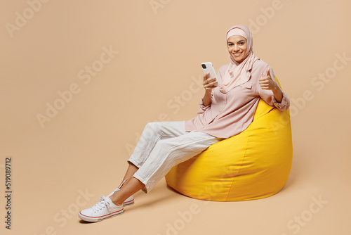 Full body young arabian asian muslim woman she wear abaya hijab pink clothes sit in bag chair use mobile cell phone show thumb up isolated on plain beige background People uae islam religious concept