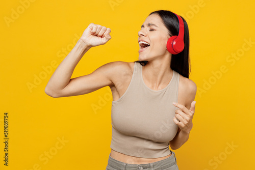 Young singer fun happy latin woman 30s she wear basic beige tank shirt headphones listen to music sing song in microphone isolated on plain yellow backround studio portrait. People lifestyle concept. © ViDi Studio