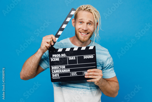 Young happy blond man with dreadlocks 20s he wear white t-shirt holding classic black film making clapperboard isolated on plain pastel light blue background studio portrait. People lifestyle concept.