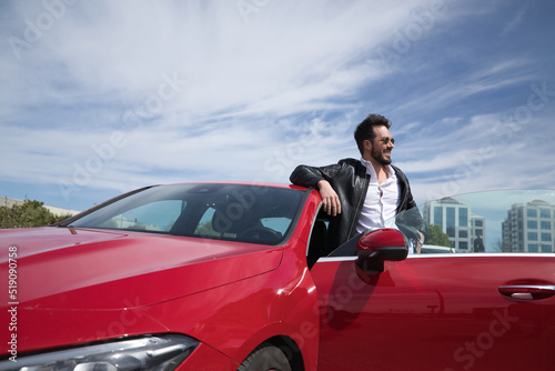 Handsome young man with beard, sunglasses, leather jacket and white shirt, leaning on the roof of his red sports car, very smiling. Concept beauty, fashion, trend, luxury, motor, sports.
