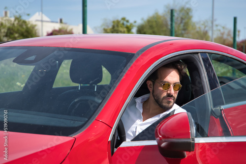 Handsome young man with beard, sunglasses and white shirt, inside his red sports car. Concept beauty, fashion, trend, luxury, motor, sports, winner.