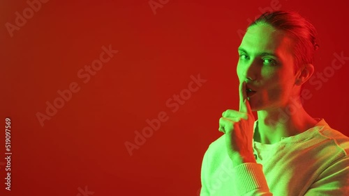 Man secret. Hush gesture. Intrigue conspiracy. Keep quiet. Green neon light smiling guy showing shh finger isolated on red color copy space advertising background. photo