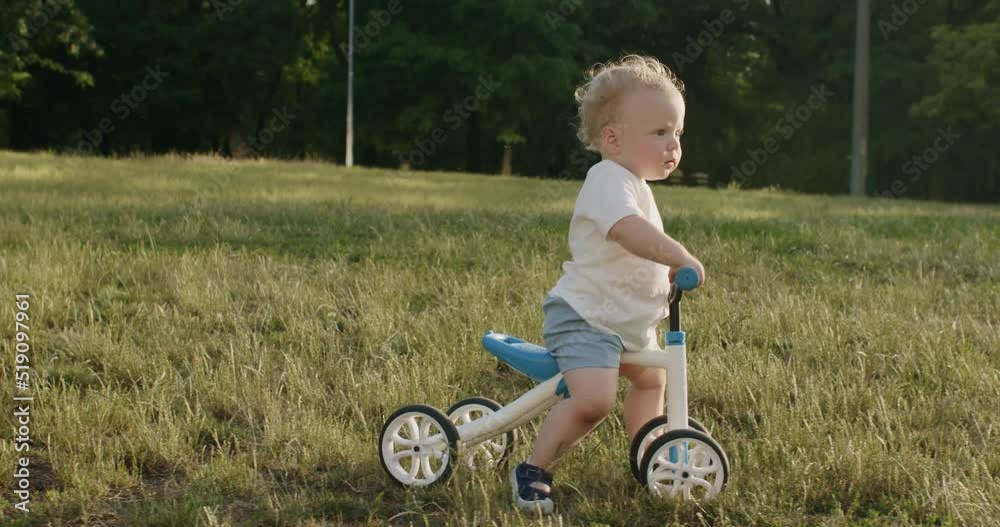 Cute blond toddler boy on kids bicycle points finger into sky. Little boy rides small bike on grass field in public park on sunny day Cute blond toddler boy on kids bicycle points finger into sky. Little boy rides small bike on grass field in public park on sunny day