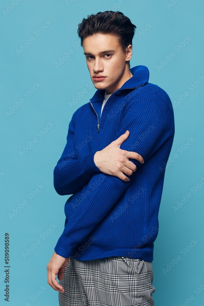 Close-up photo. a young and slender man with thick black hair standing on a blue background in a blue zip-up sweater with a white T-shirt under it. Close-up photo. a young and slender man with thick black hair standing on a blue background in a blue zip-up sweater with a white T-shirt under it.