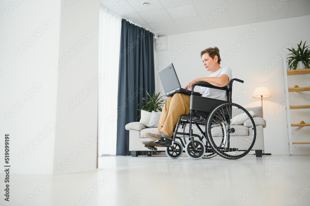 Middle age grey-haired disabled woman having video call sitting on wheelchair at home. Middle age grey-haired disabled woman having video call sitting on wheelchair at home.