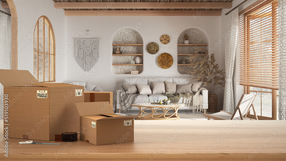 Obraz premium Wooden table, desk or shelf with stack of cardboard boxes over blurred view of bsohemian living room in boho style, bohemian interior design, moving house concept with copy space