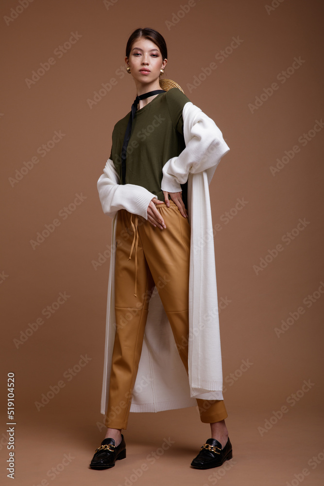 Fashion photo of a beautiful elegant young asian woman in a pretty beige leather pants, white long cardigan, green t-shirt, belt posing over brown coffee background. Studio Shot. Portrait