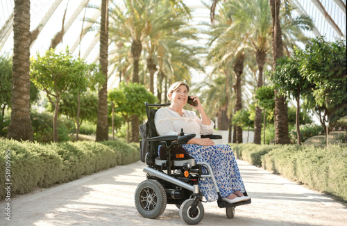 a smiling woman sitting in an electric wheelchair enjoys a sunny day in the garden of the city park talking on her mobile phone