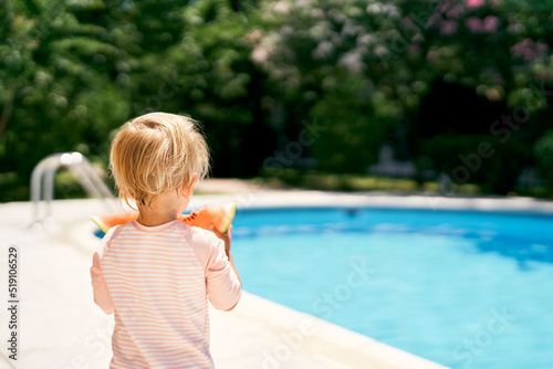 Little baby stands by the pool, holding a watermelon in his hands. Back view