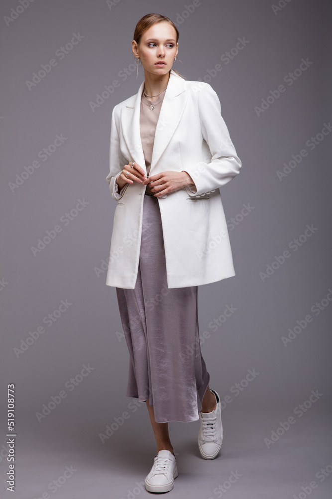Fashion photo of a beautiful elegant young woman in a pretty white jacket, long metallic silver platinum skirt, sneakers posing gray background. Studio Shot. Portrait Fashion photo of a beautiful elegant young woman in a pretty white jacket, long metallic silver platinum skirt, sneakers posing gray background. Studio Shot. Portrait
